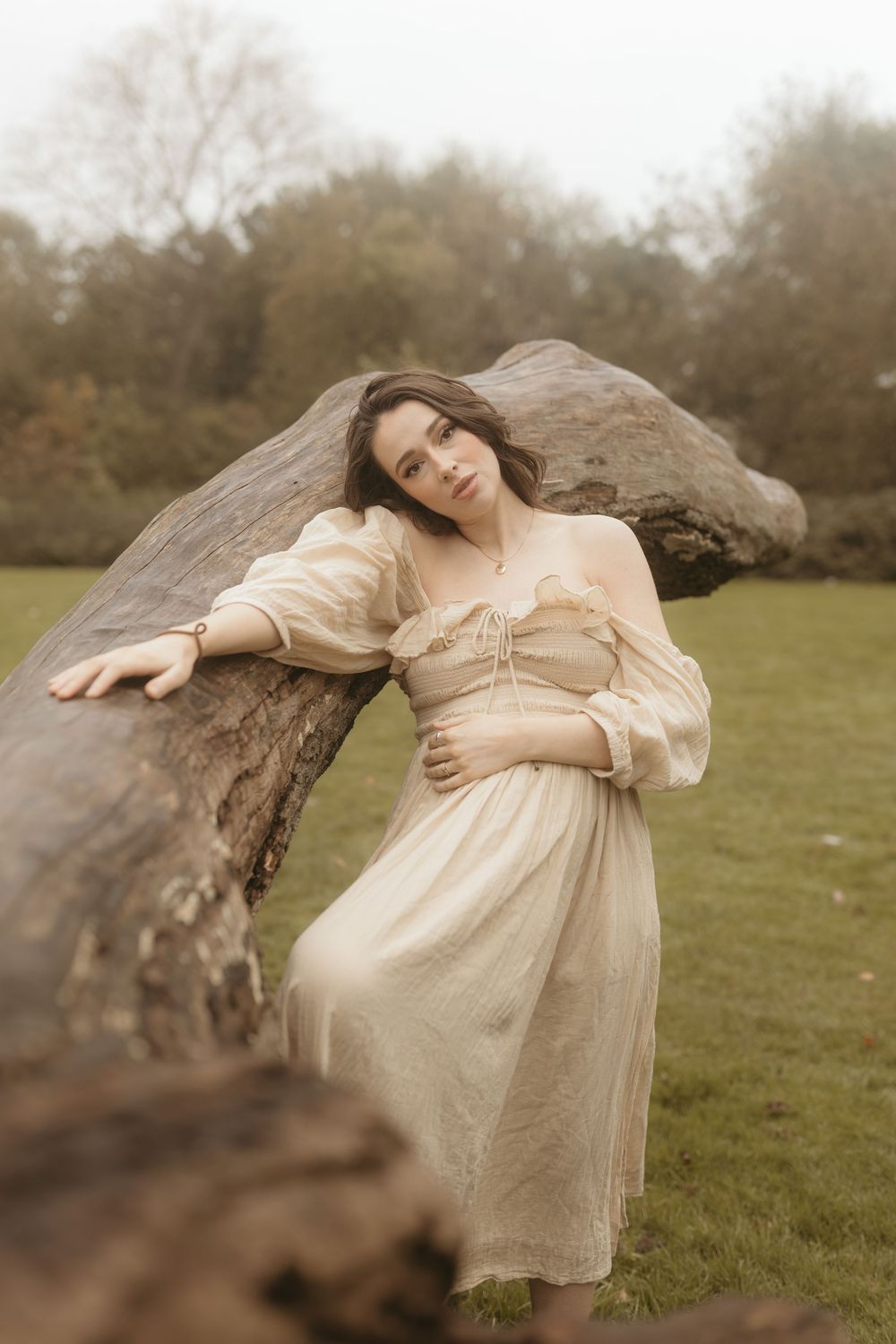A romantic ethereal portrait in a cream dress leaning against a fallen tree trunk in a dreamy outdoor setting.