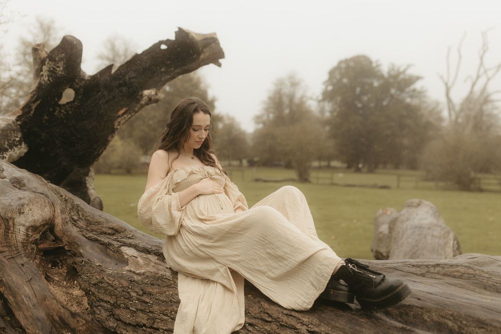 A dreamy countryside portrait showing a person in a flowing cream dress reclining on a fallen tree trunk on a misty day.