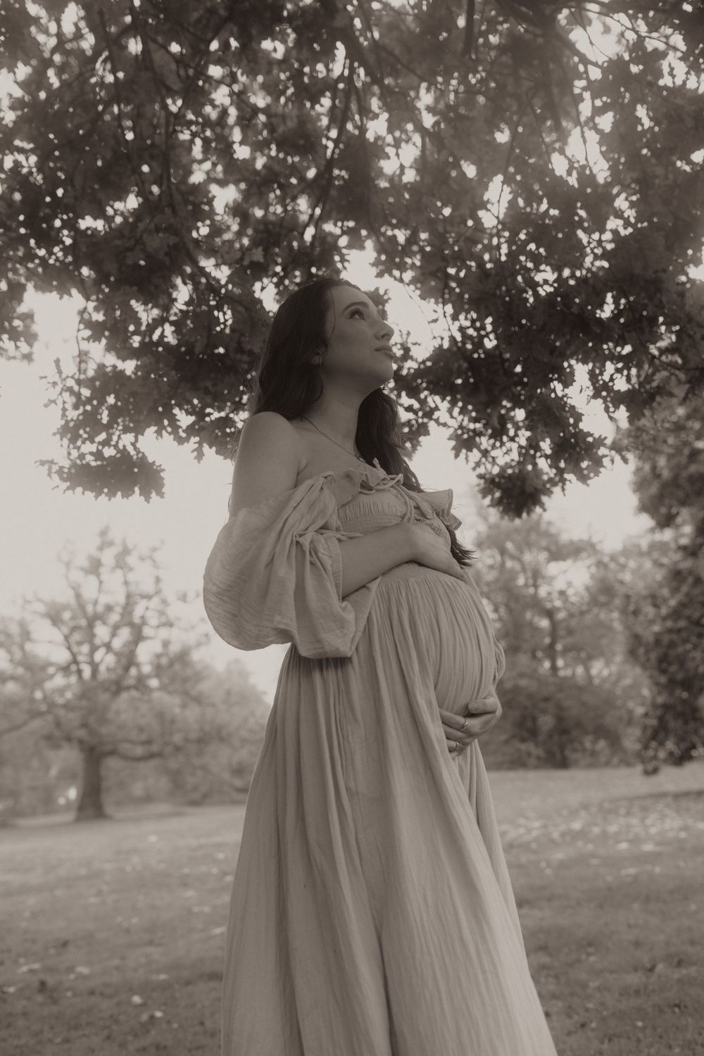 A dreamy black and white maternity photo shows an expectant mother in a flowy dress standing beneath tree branches.