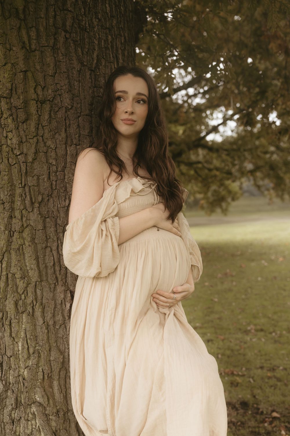 An expectant woman in a flowing cream dress poses gently against a tree trunk in a dreamy outdoor setting.