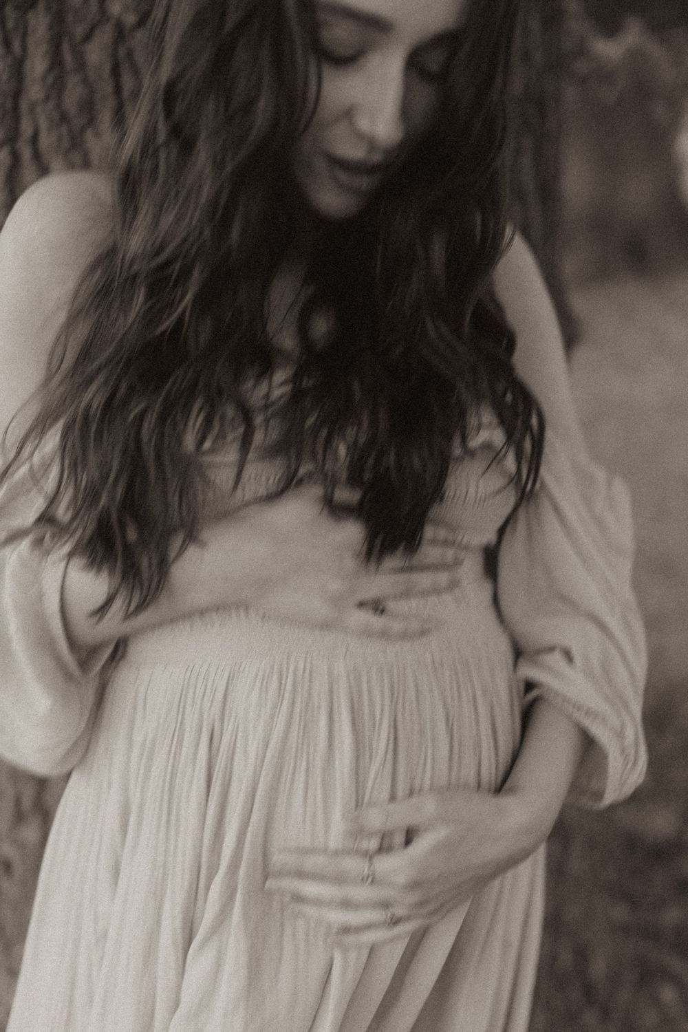 Black and white portrait of a pregnant woman in a flowy dress with long wavy hair looking downward.