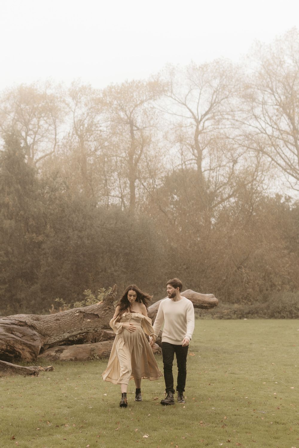 A romantic couple walks together in a misty park setting near a fallen tree trunk.