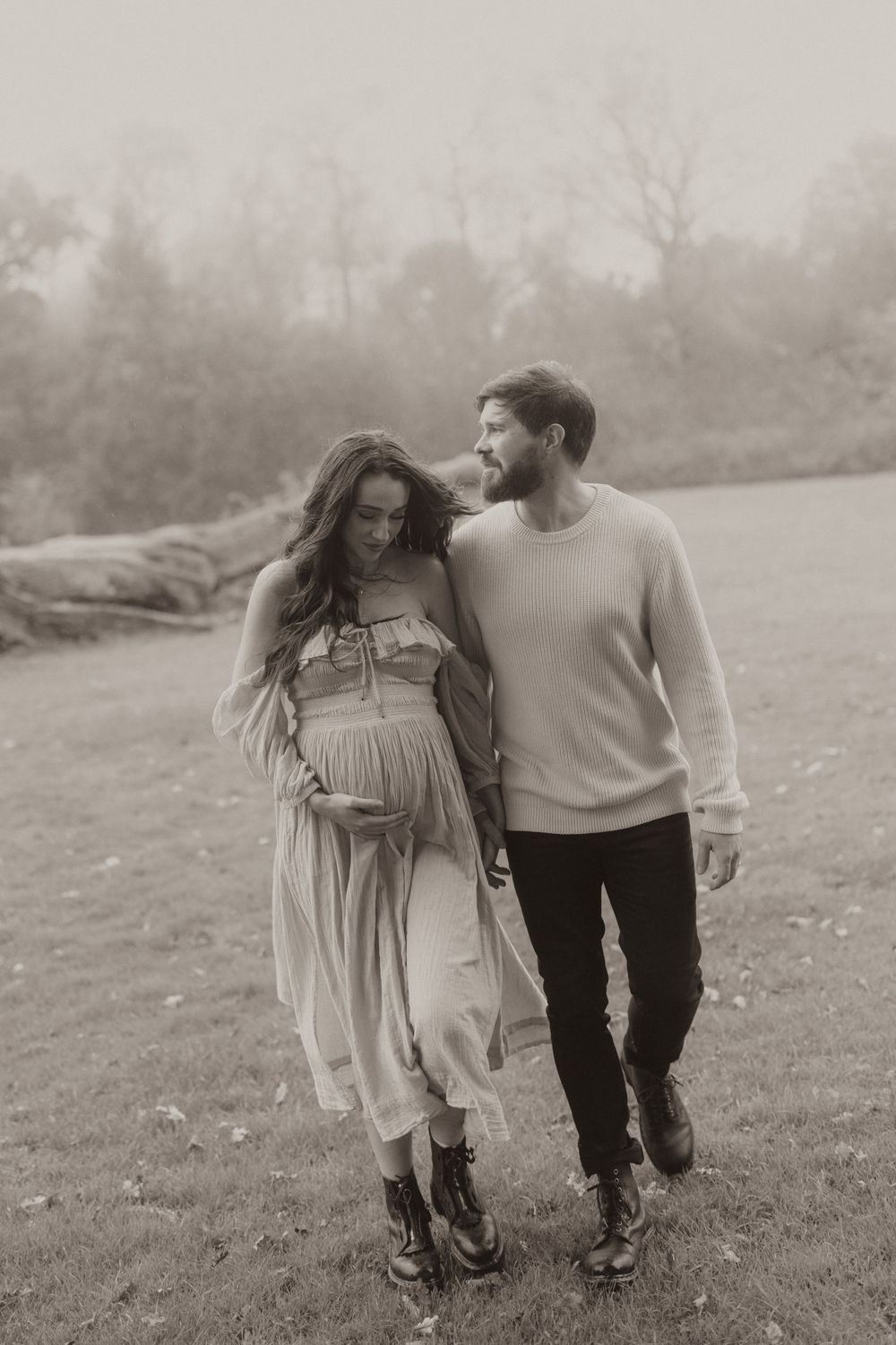 A romantic couple walks together on a misty field, with the pregnant figure wearing a flowing dress and companion in sweater.