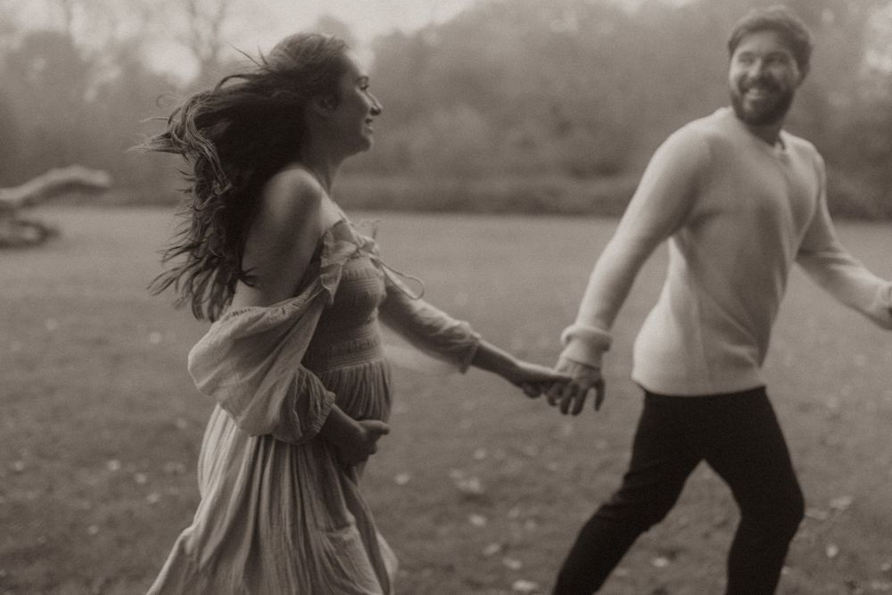 Couple in black and white holding hands and running playfully through a misty field with flowing dress and casual attire.