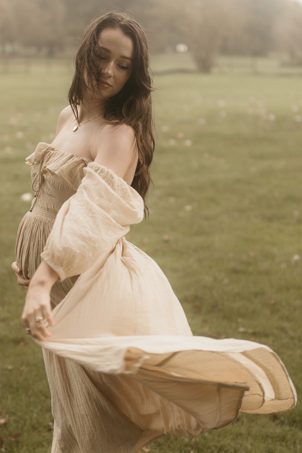 Woman in flowing beige vintage-style dress twirls gracefully in a misty green field at golden hour.
