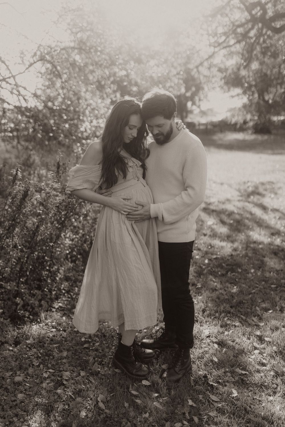 An expectant couple shares a tender moment outdoors while wearing cozy sweaters on a misty afternoon.