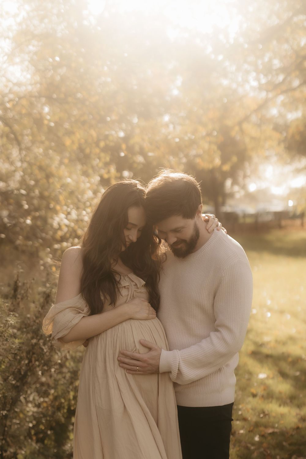A tender maternity moment captured in golden light as a couple embraces amidst blooming spring trees.