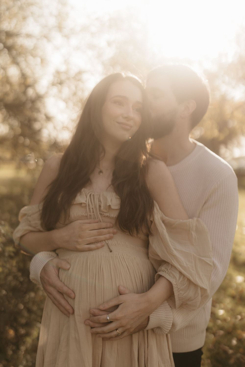 A romantic sunlit maternity photo captures a tender moment between a couple in light colored clothing.