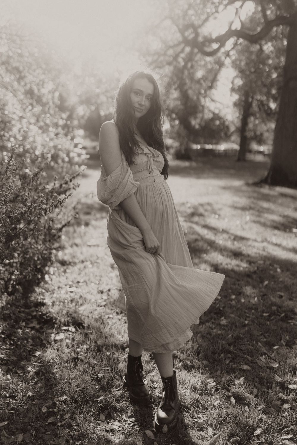 A vintage-style maternity portrait shows a woman in a flowing dress posing outdoors in dappled sunlight.