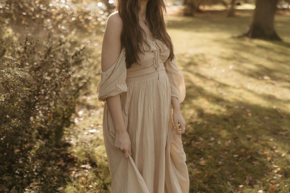 Woman wearing romantic off-shoulder cream dress poses in dappled sunlight among trees in a dreamy garden setting.