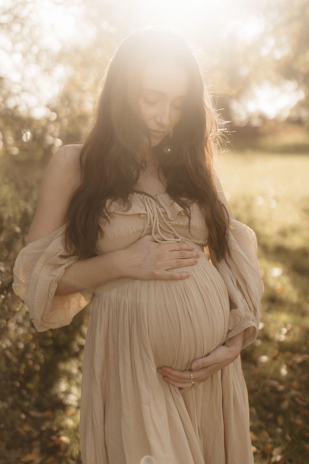 A pregnant woman in a flowing white dress stands in a sunlit field at golden hour, gently cradling her baby bump.