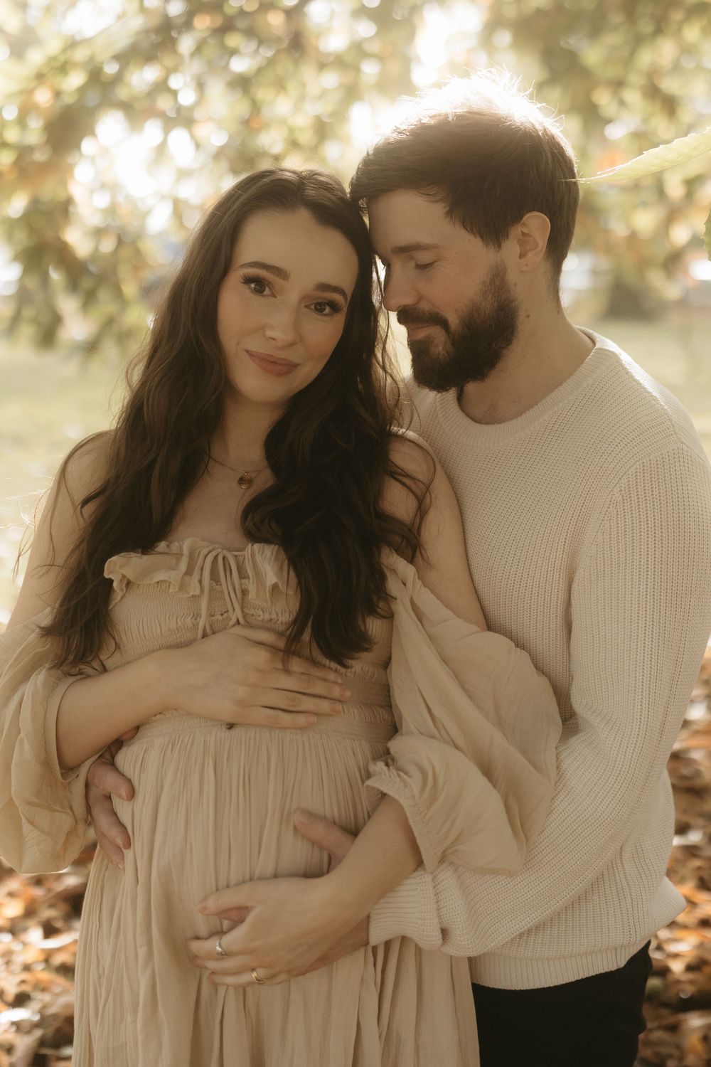 Couple in romantic autumn maternity photo shoot with beige dress and white sweater sharing intimate moment in nature.