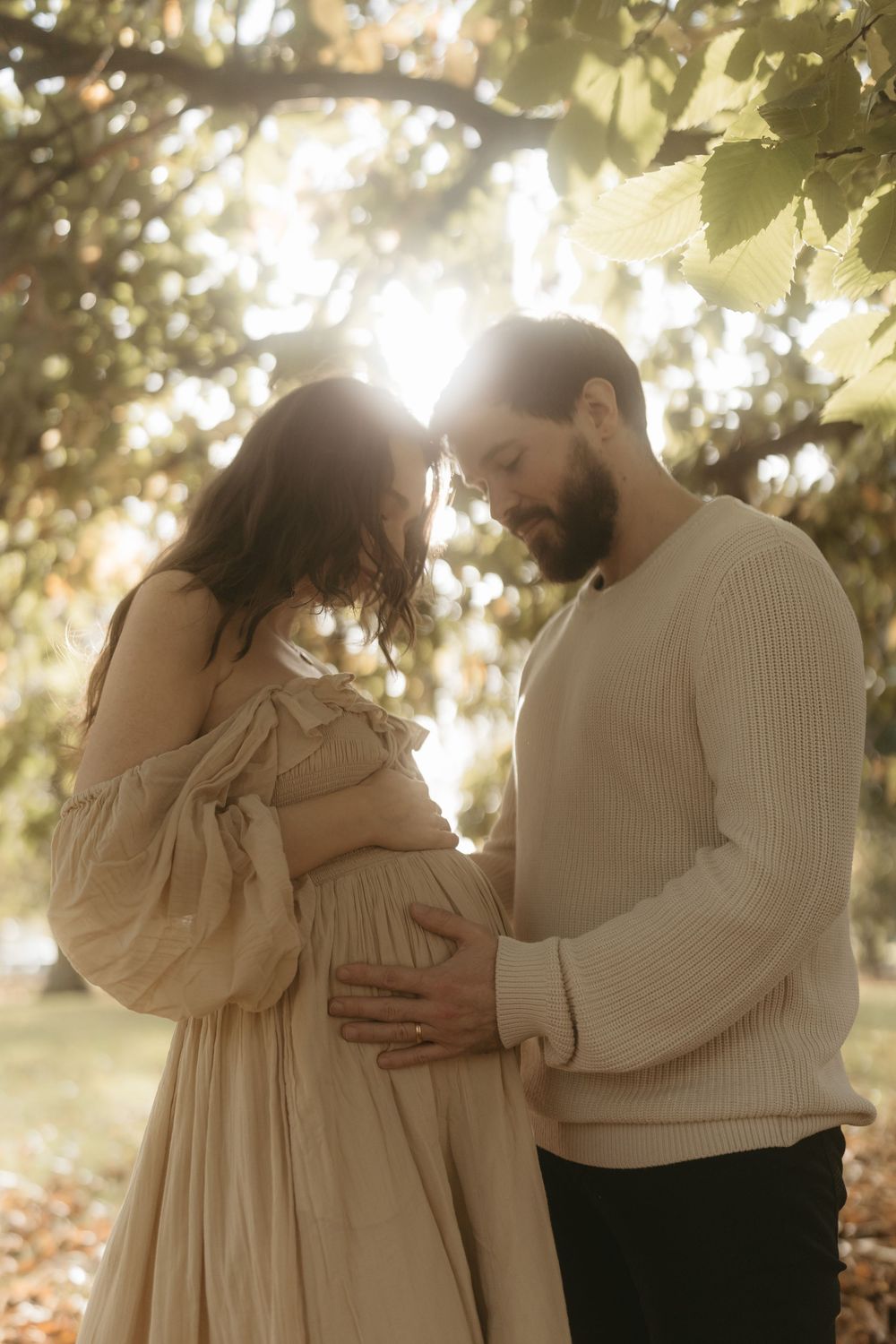 Romantic couple shares intimate moment outdoors as sunlight filters through trees, showcasing a loving pregnancy photo.