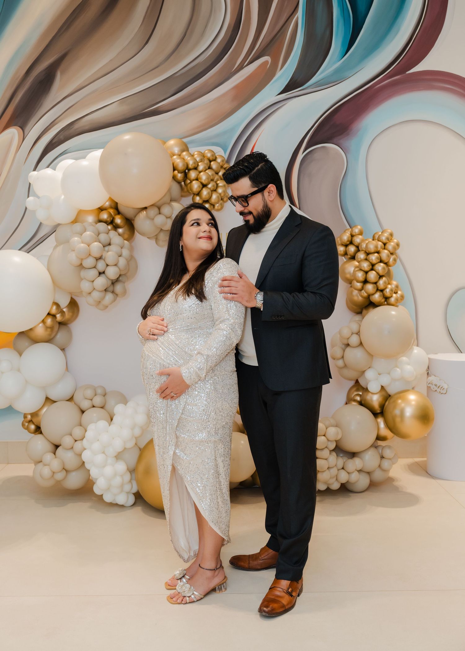 Elegant couple poses in formal attire against a luxurious balloon arch backdrop with gold and white decorations.
