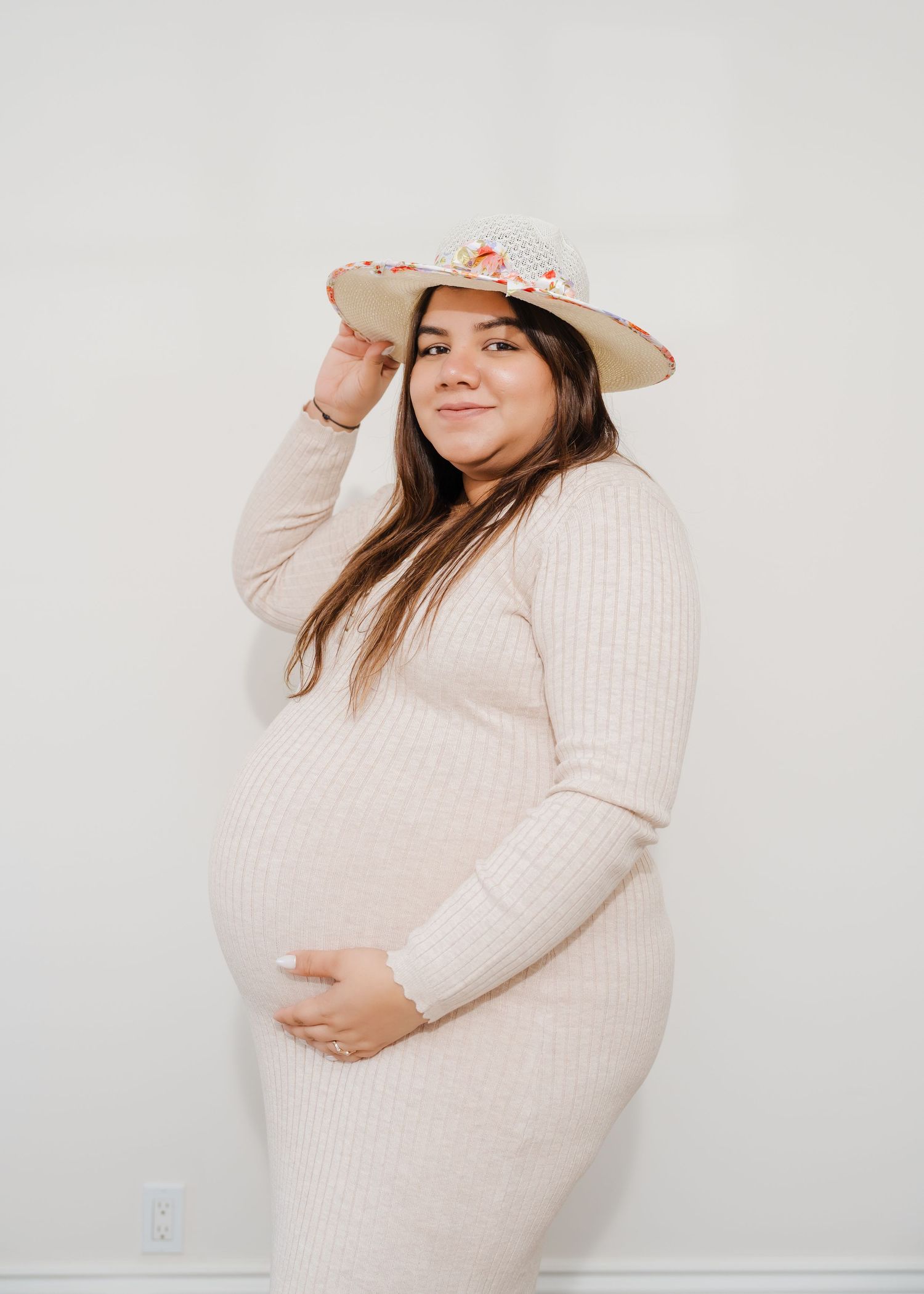 A pregnant person in a cozy cream sweater dress and floral hat poses against a white wall.