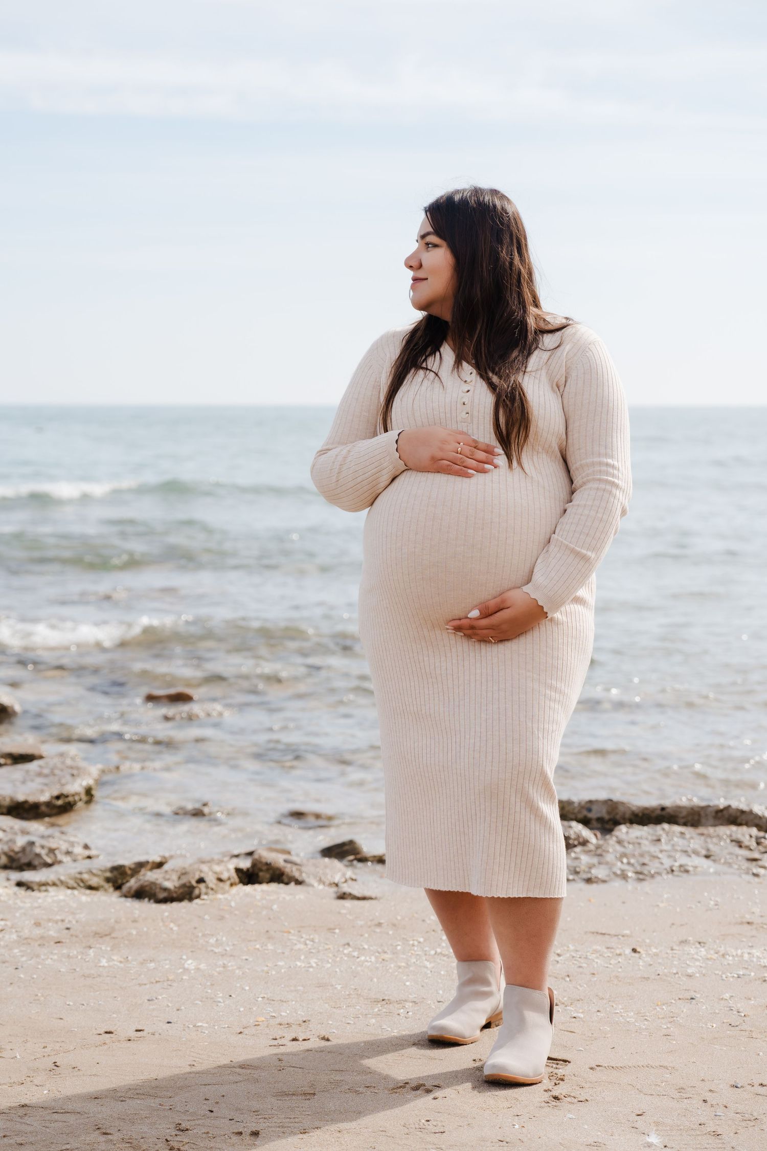 Expectant person in cream knit dress poses thoughtfully on rocky beach shoreline in soft sunlight.
