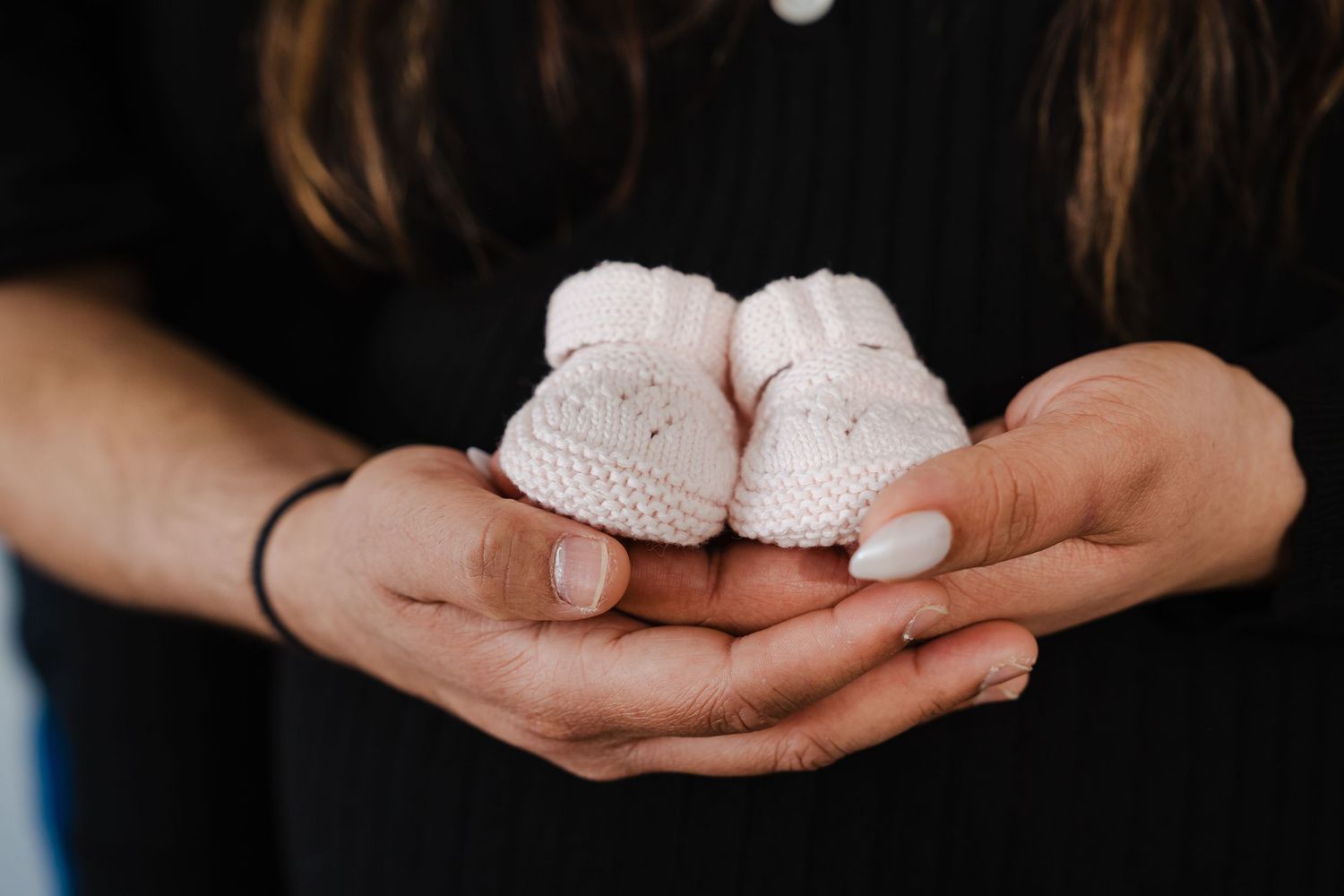 A pair of tiny pink knitted baby booties held gently in outstretched hands with white manicured nails.