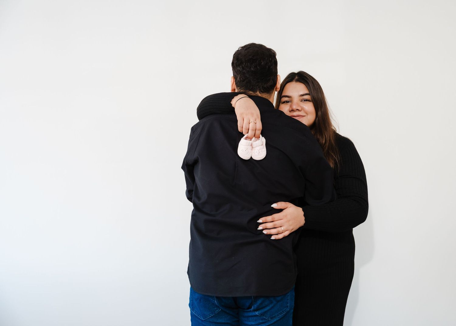 A couple embraces tenderly against a white wall wearing matching black sweaters.