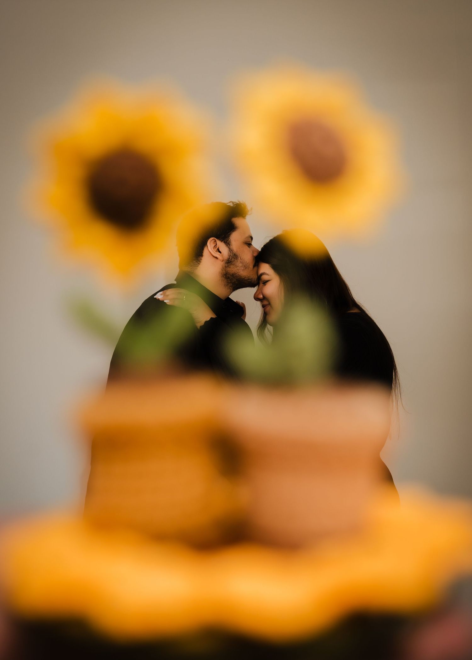 A romantic couple shares an intimate moment with sunflowers in soft focus behind them.