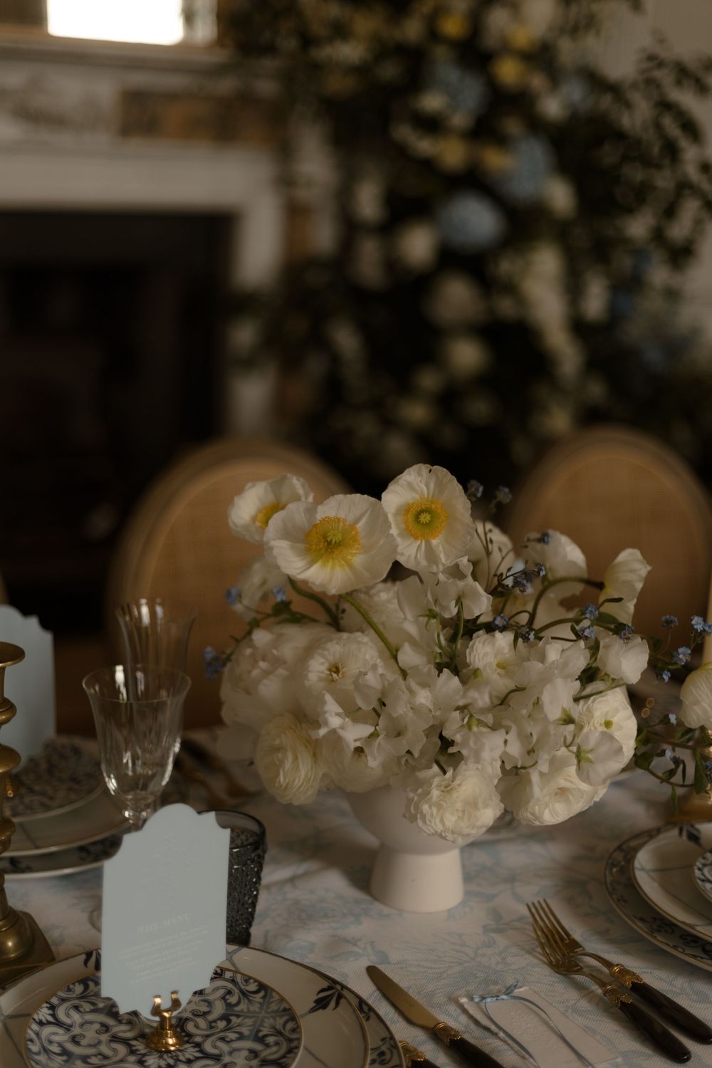 Elegant white floral centerpiece with roses and daffodils on a formal table setting with crystal glassware and marble surface.