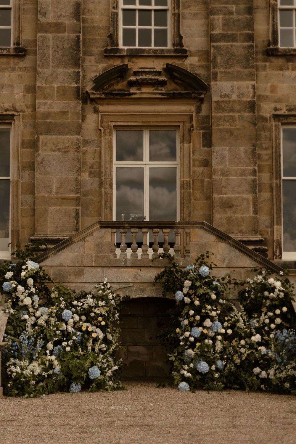 Elegant historic stone building entrance with ornate window and lush white and blue floral arrangements framing the doorway.