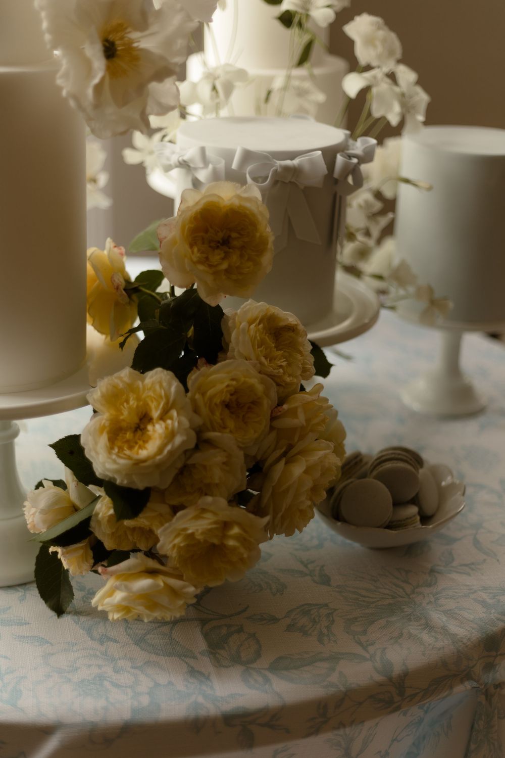Elegant table setting with pale yellow garden roses and white ceramic cake stands against a delicate patterned tablecloth.