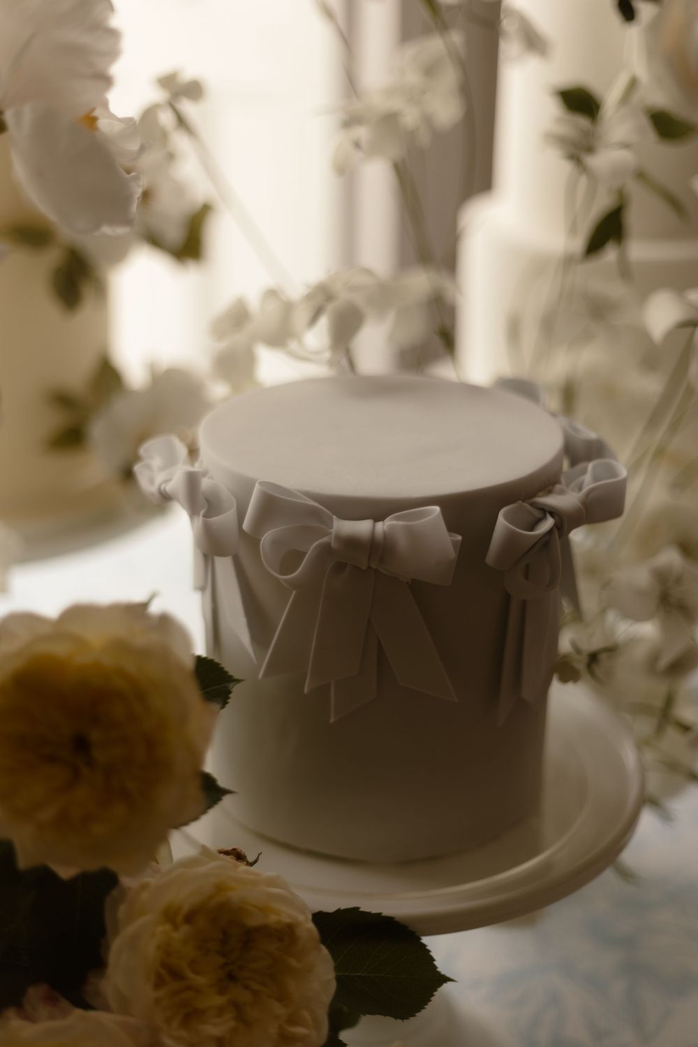 White ceramic cake top decorated with ribbons and bows sits elegantly among soft yellow roses in dreamy lighting.