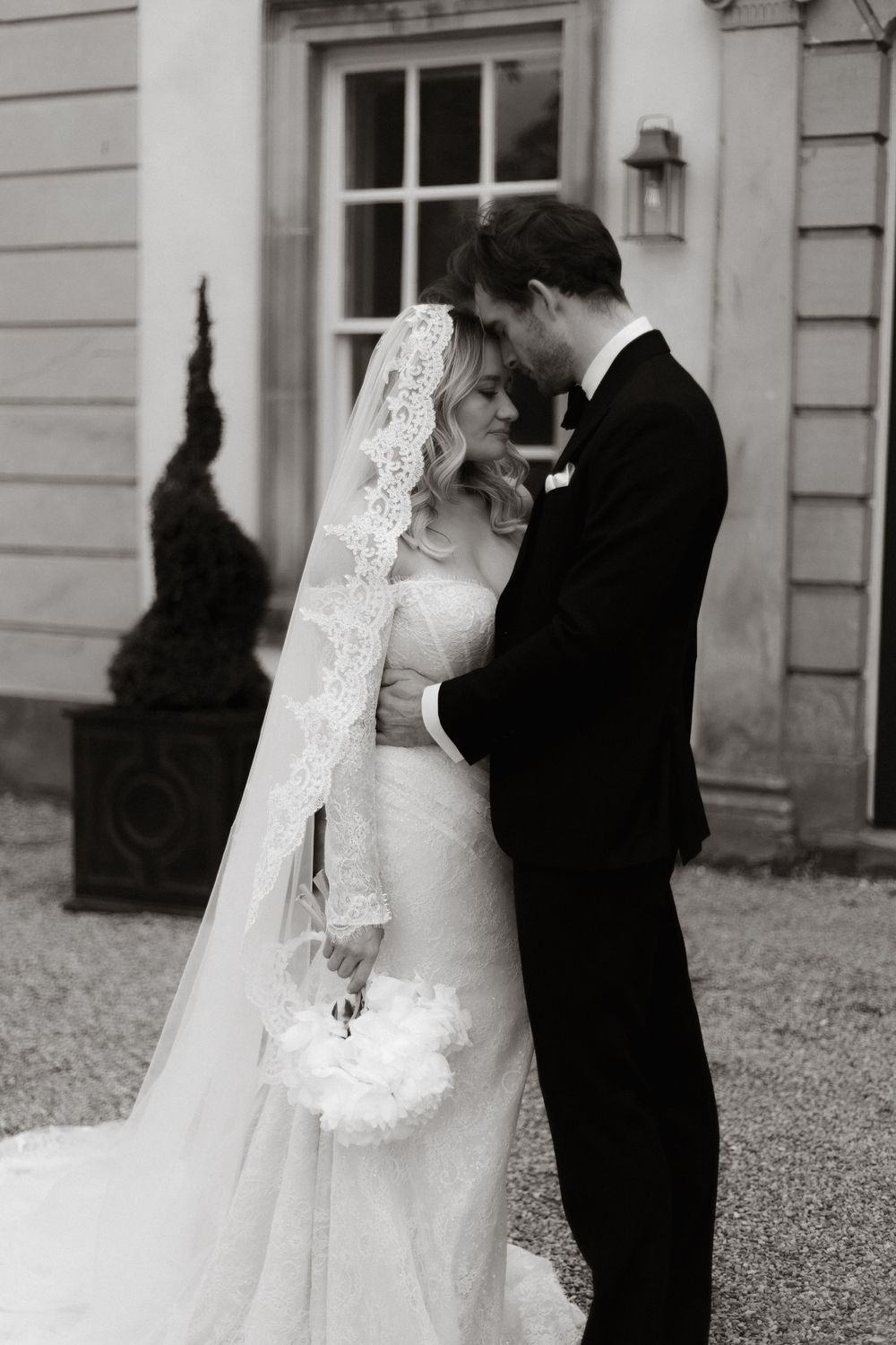 A romantic black and white wedding photograph of newlyweds sharing an intimate moment outdoors by a building entrance.