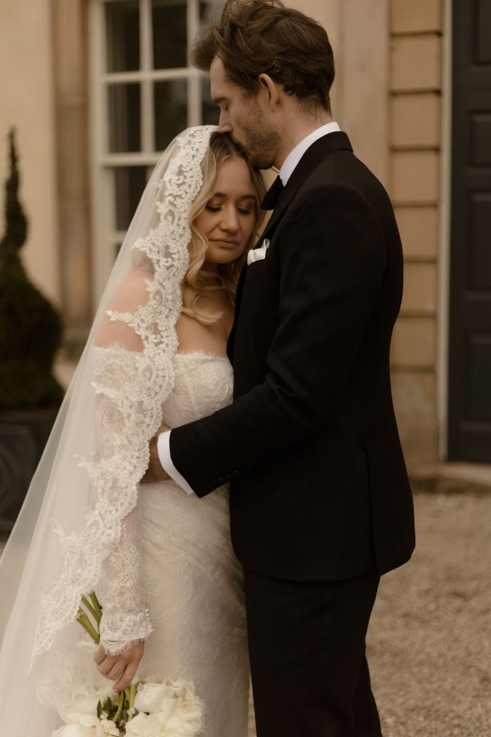 A romantic wedding moment between a couple, with the bride wearing a lace veil and white gown against an elegant building exterior.