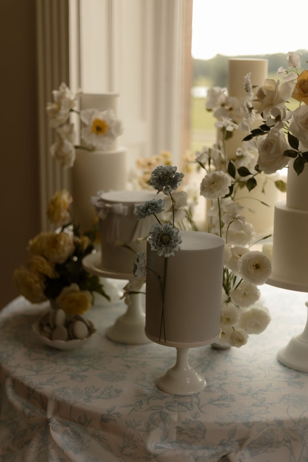 Elegant white wedding cakes adorned with yellow and white flowers on pedestal stands against a sunlit window.