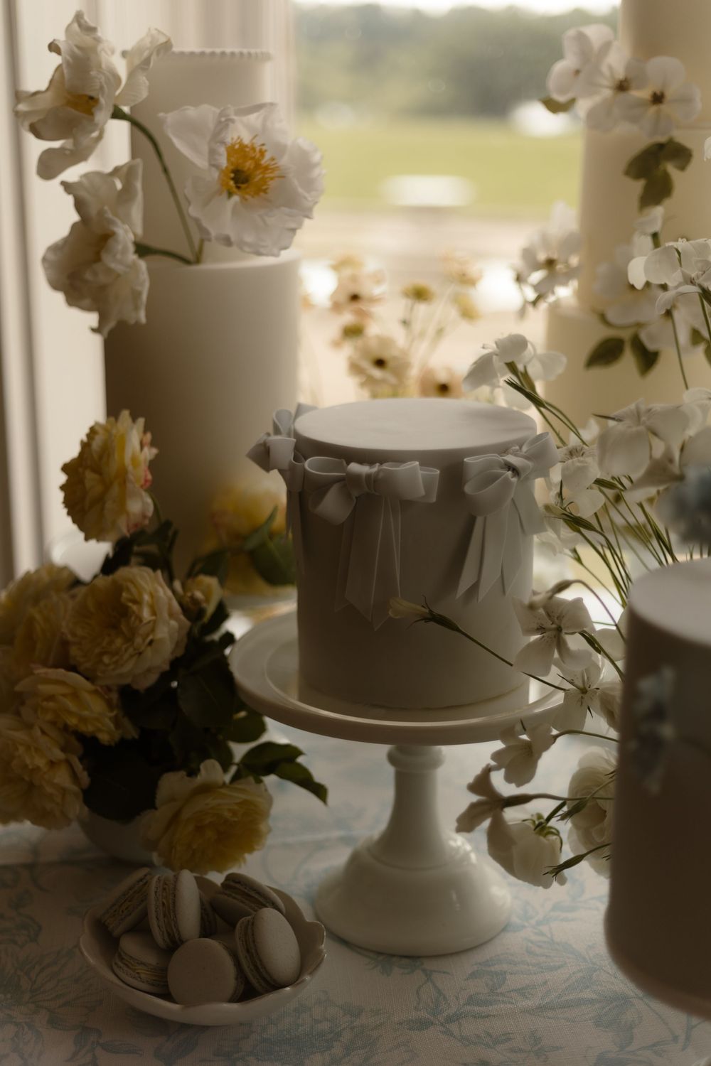 White wedding cake on pedestal surrounded by delicate flowers and macarons in a soft, dreamy setting.