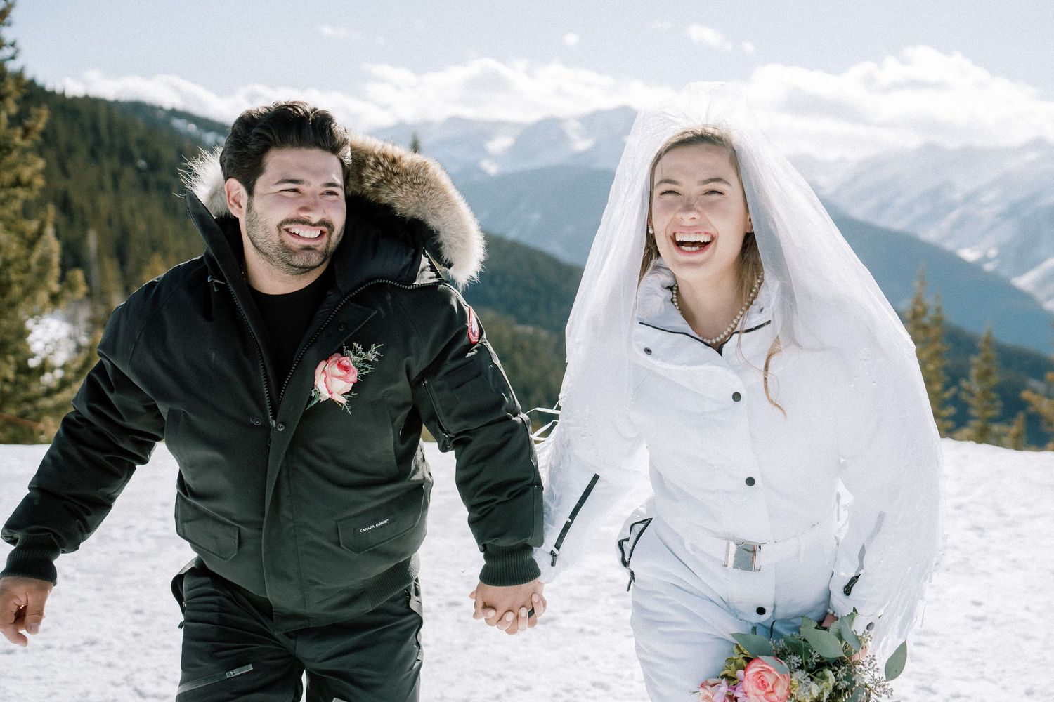 Newlyweds in winter attire run hand-in-hand through snow with mountain backdrop during winter elopement.