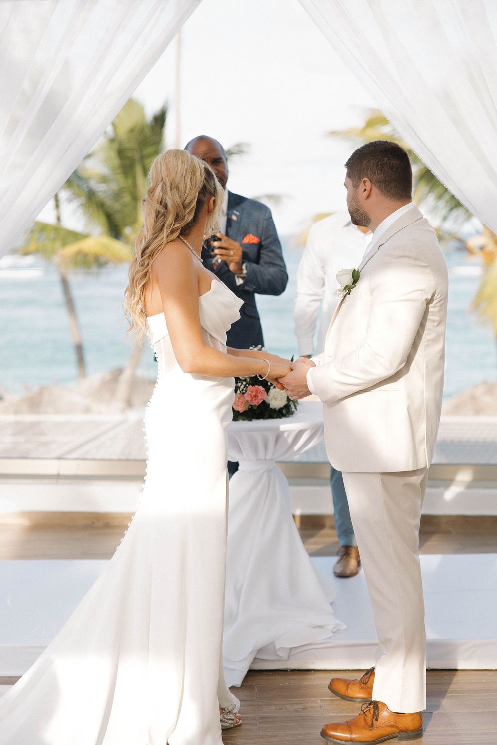 A beachside wedding ceremony takes place under a white canopy with palm trees and ocean views in the background.