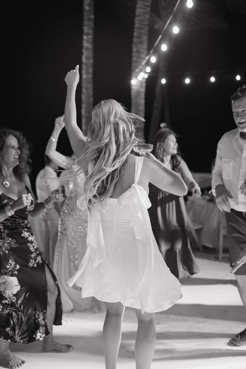 Black and white photo of wedding reception dancing under string lights with flowy white dress twirling on dance floor.