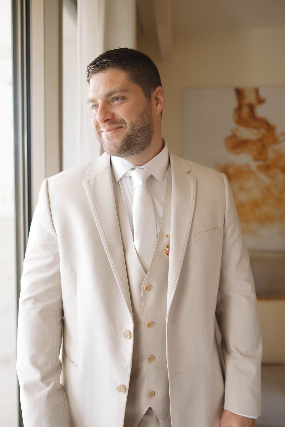 A groom in a classic cream-colored three-piece suit stands by a window, smiling while getting ready for his wedding day.