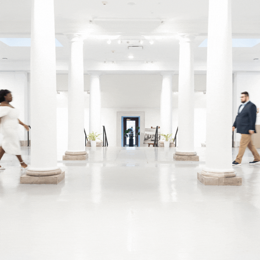 Two figures crossing through a minimalist white hallway with columns and art displays in a modern museum interior.