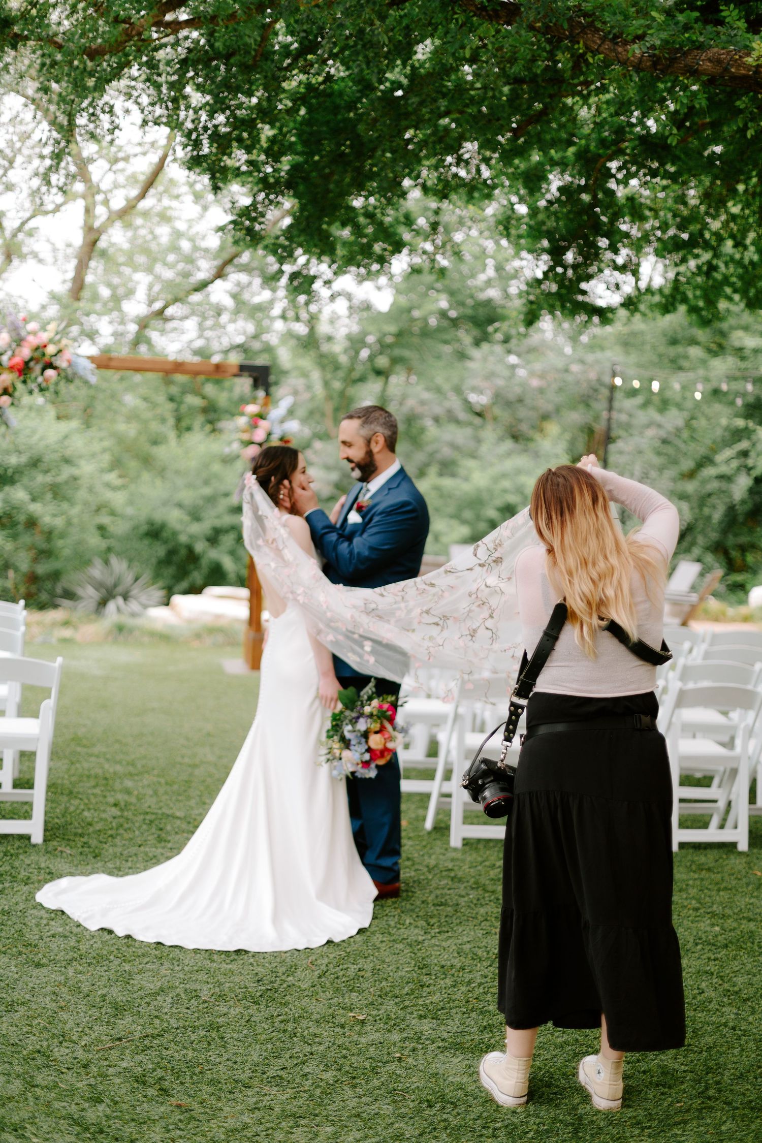 Wedding photographer in black dress capturing a couple during their outdoor garden ceremony with white chairs setup.