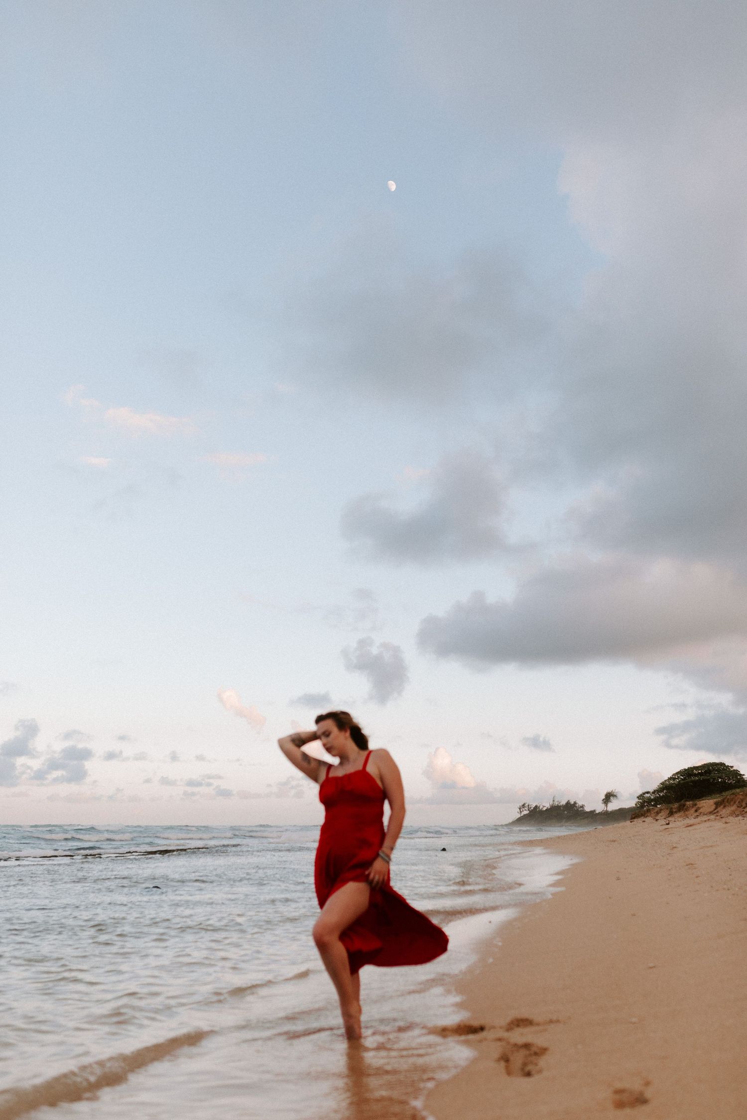Woman in flowing red dress dancing on beach at sunset with ocean waves in background.