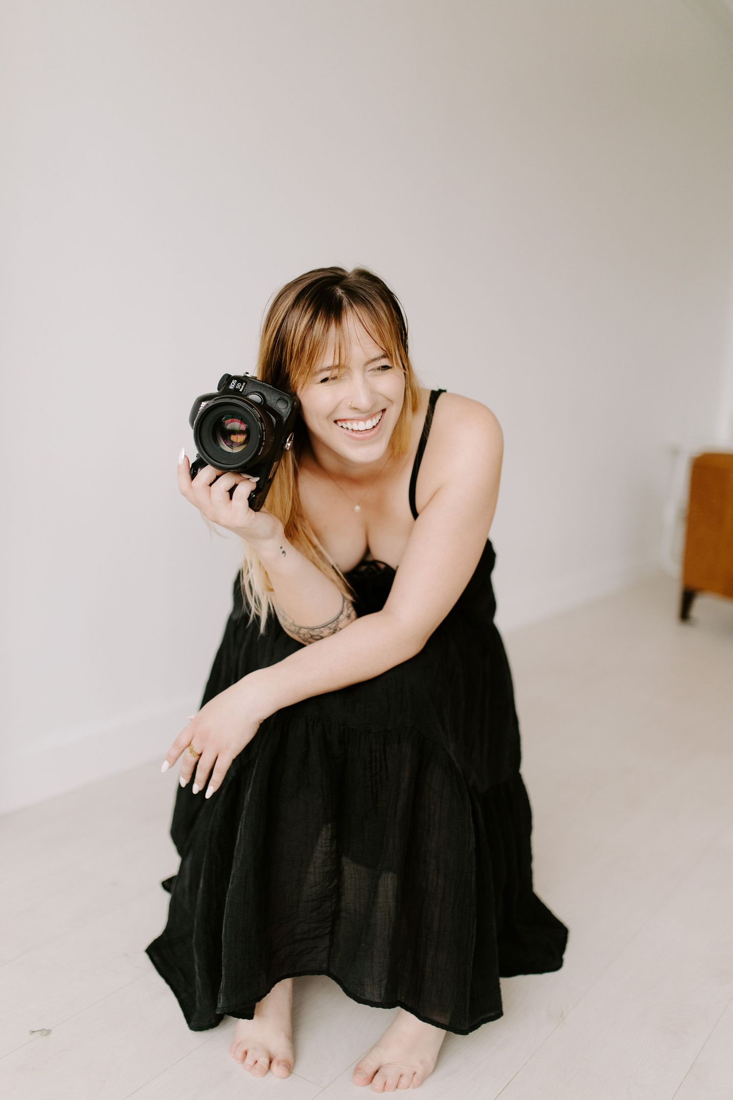 Portrait of photographer holding camera while sitting in black dress against white backdrop.