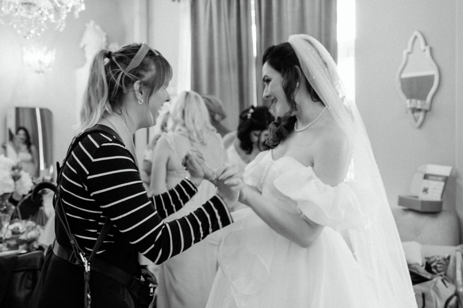 Intimate black and white moment shows bride getting ready with helper in striped shirt.