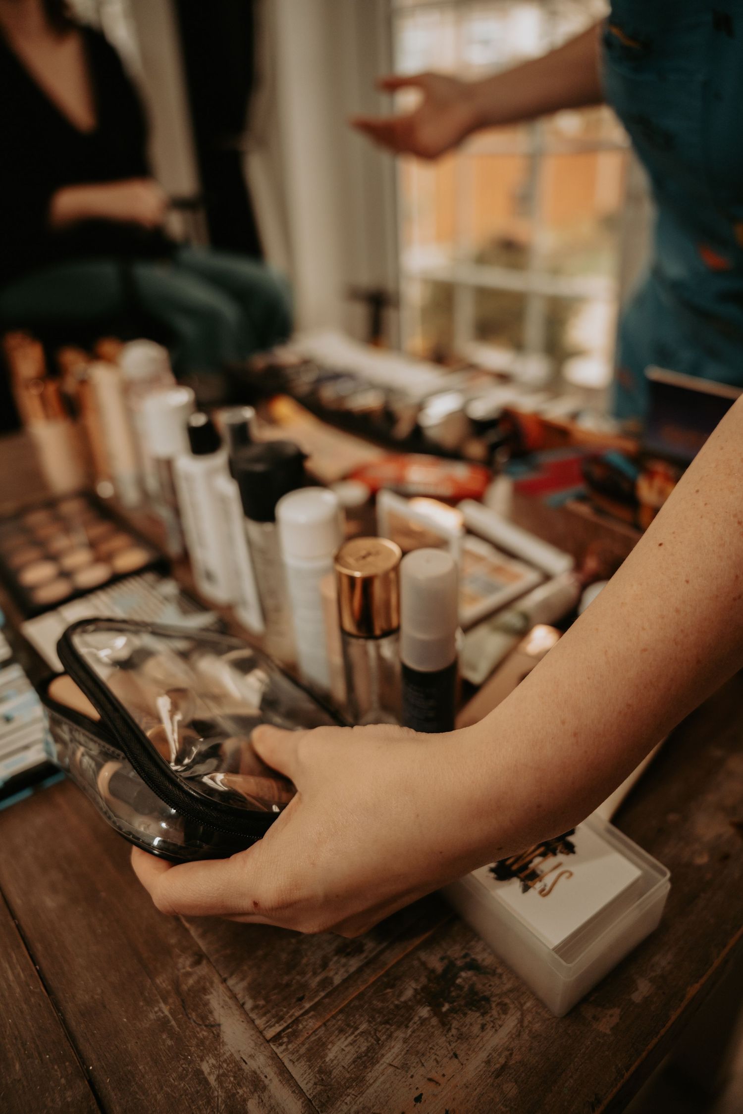 Makeup artist preparing cosmetics and beauty products on wooden table with eyeshadow palettes and bottles displayed.