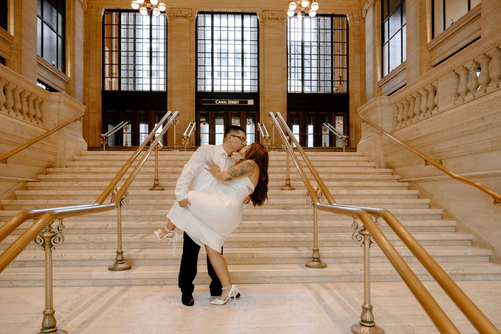 A romantic couple dips and embraces on an elegant marble staircase with ornate gold railings and tall arched windows. Wedding Photography