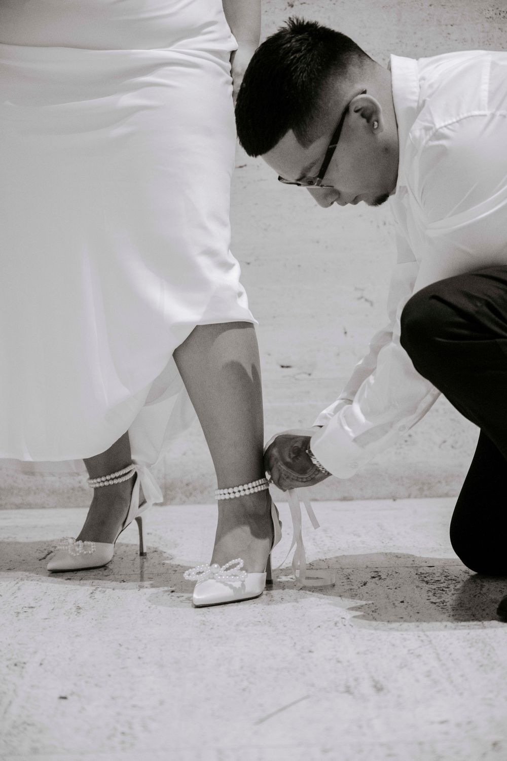 A person in black crouches to adjust an elegant white bridal shoe adorned with pearl ankle straps. Wedding Photography