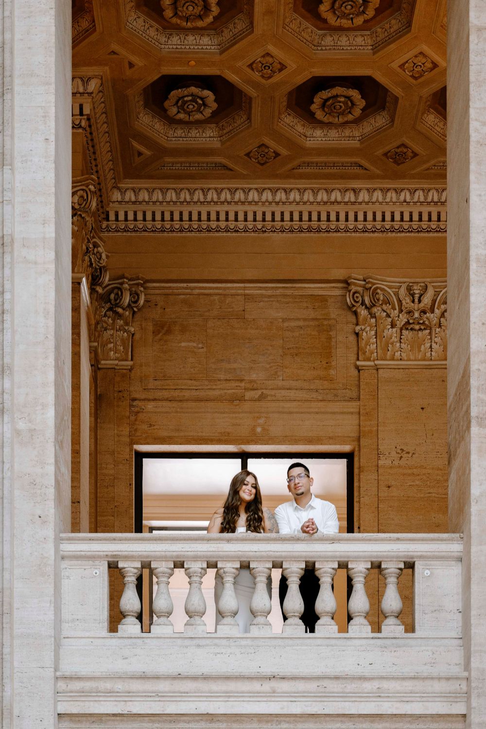 An ornate architectural interior with stone balustrade & decorative coffered ceiling features two people standing at a balcony.