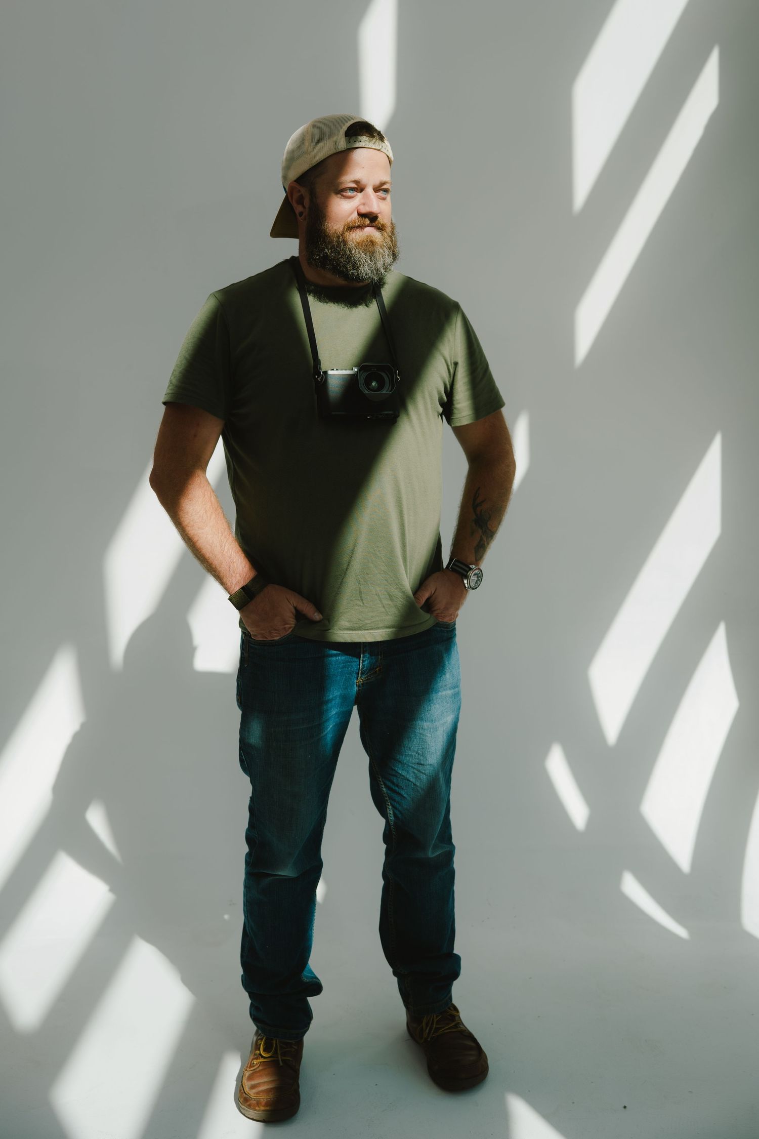 A person wearing a green t-shirt, beanie hat, and jeans stands against a white wall with dramatic diagonal shadows.