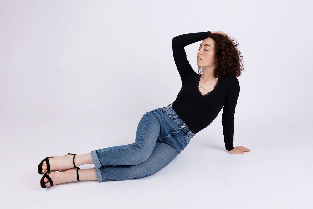 A relaxed casual pose in a black long-sleeve top and blue jeans with strappy sandals on a white background.