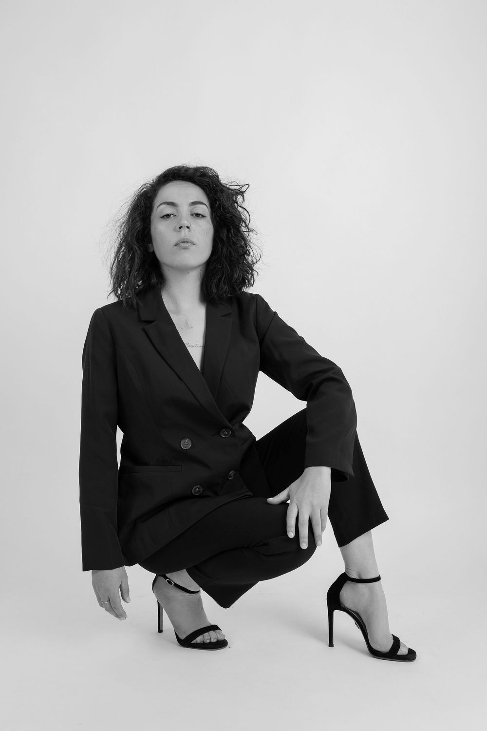 Black and white fashion photo of person in dark blazer suit and heels crouching against white backdrop.