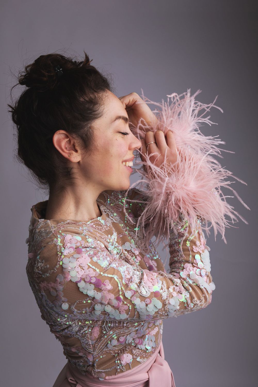 A joyful pose in a pink feathered and sequined sheer dress with floral embellishments against a gray backdrop.