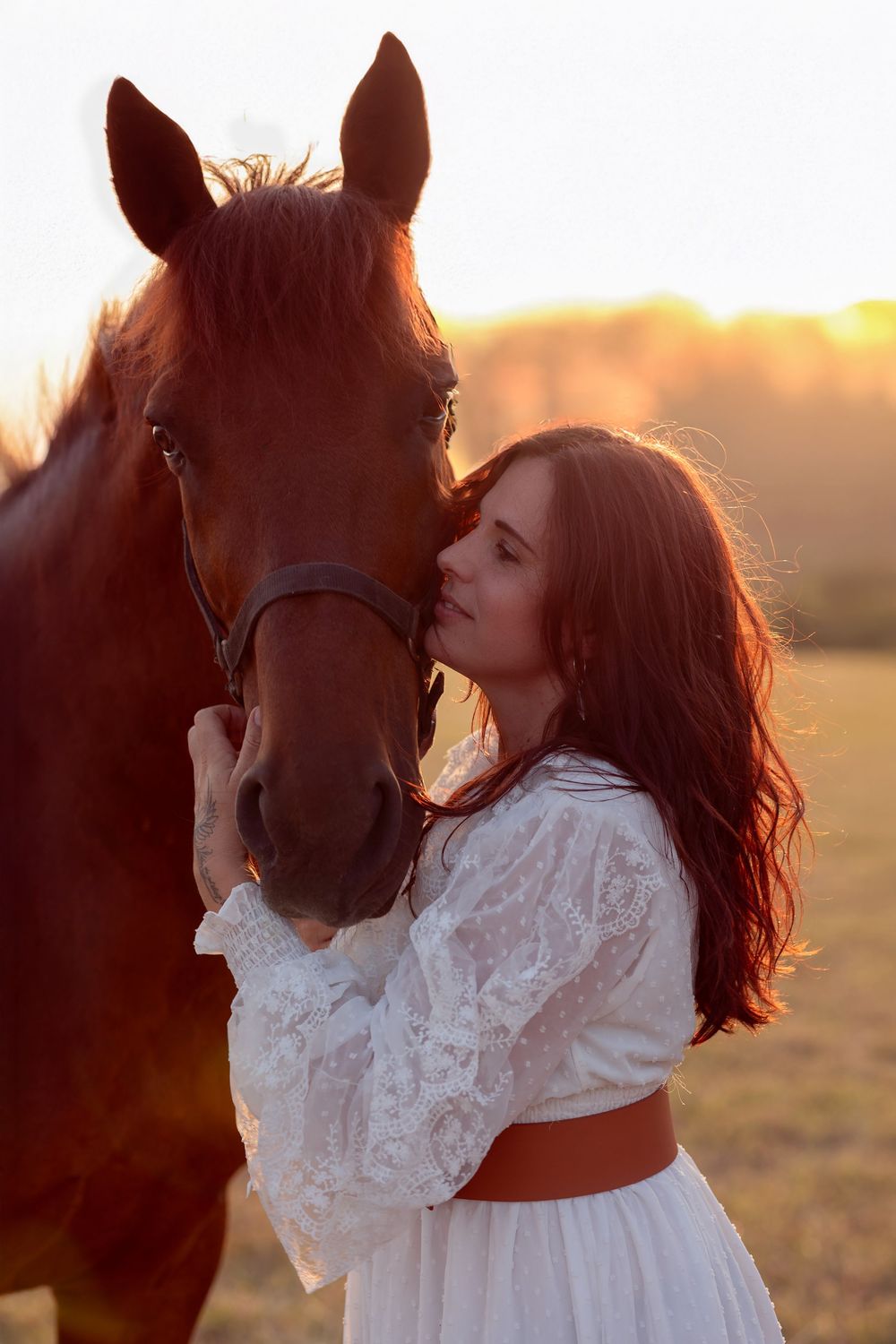 Woman in white lace dress embraces brown horse in golden sunset field.