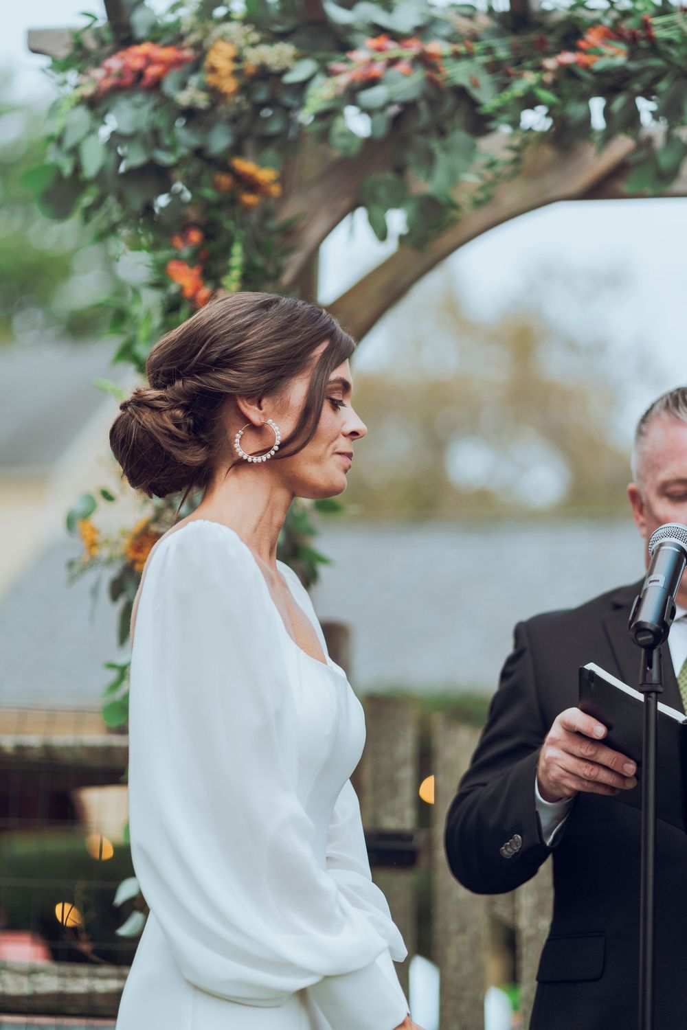 A bride hears vows from her soon to be husband in a white wedding dress with a flowery backdrop, capturing a romantic wedding scene